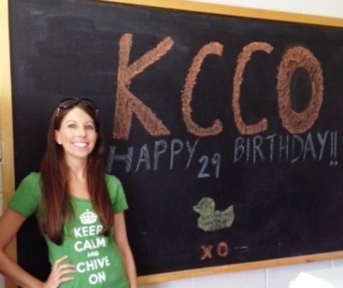 Pretty brunette in green KCCO tee poses near chalkboard with birthday message to KCCO