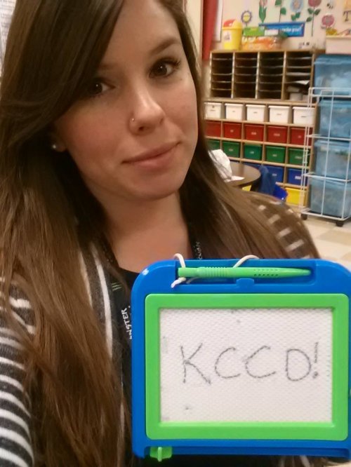 Brunette teacher with KCCO sign takes selfie in black top and black/white striped jacket