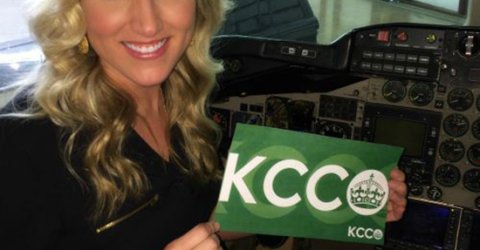 Blonde in black top smiles and poses with green KCCO sign