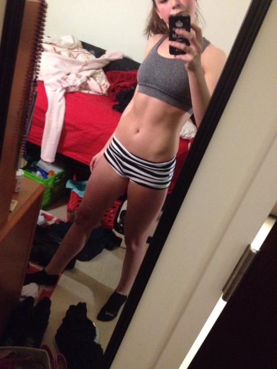 Blonde with perky boobs, flat abs, slender legs, and slim sexy hot body takes selfie in greyish-black sports bra and black/white striped short shorts