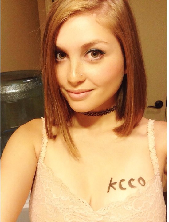 Girl with short hair wearing a choker and pink lace with KCCO written on her boobs