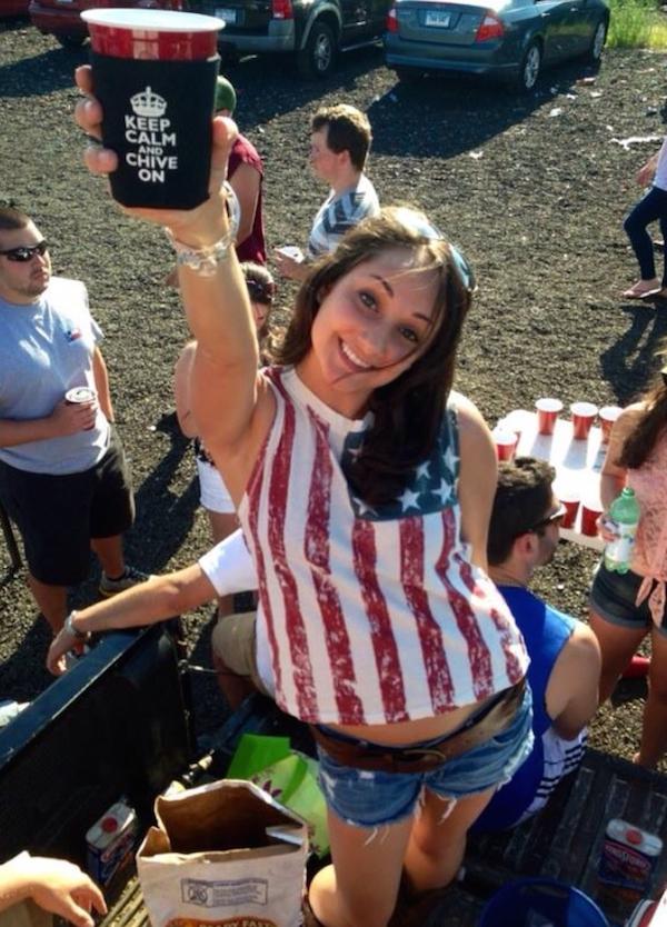 Pretty brunette with slim sexy hot body in stars and stripes top and blue denim shorts poses with black KCCO beer cup