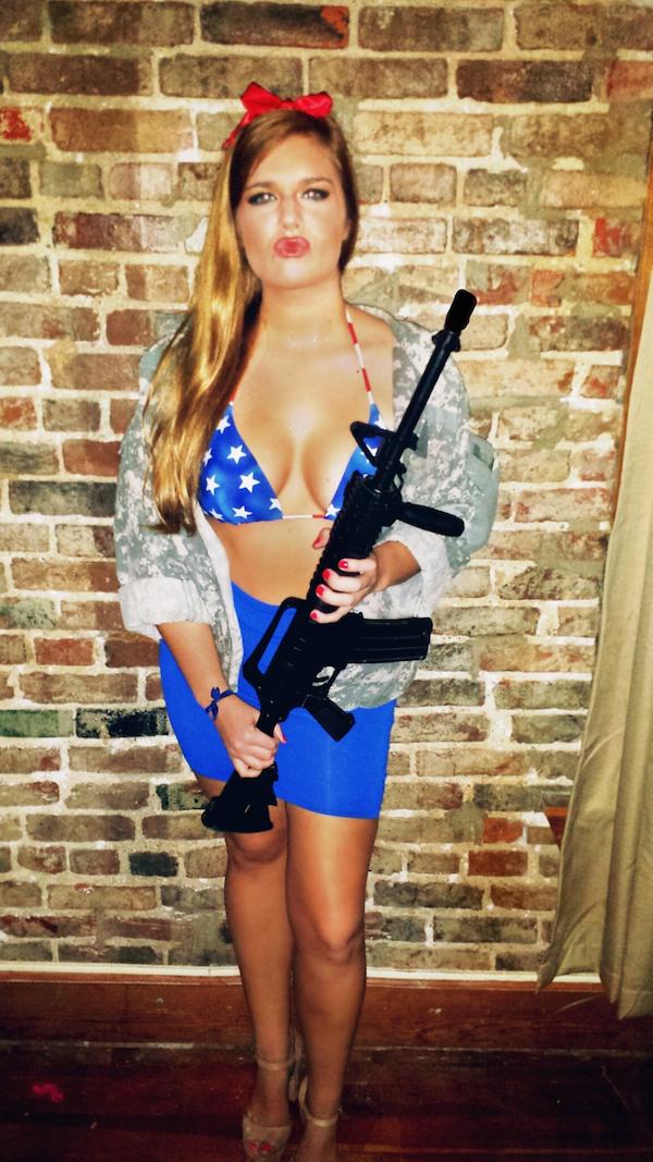Light-eyed blonde with red juicy lips, perky boobs, and hot voluptuous curvy body poses with gun in blue tight mini skirt, stars and stripes bikini top, and grey/white jacket