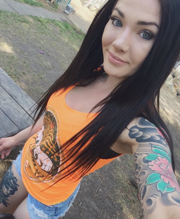 Beautiful gray eyed girl taking a designer tattoo selfie to showcase her colorful designer hand and thigh tattoo in orange top and denim shorts
