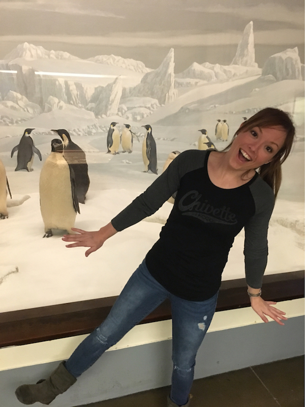 Cute girl in black top with Chivette logo, ragged jeans, brown long boots looking funny with penguins in polar background