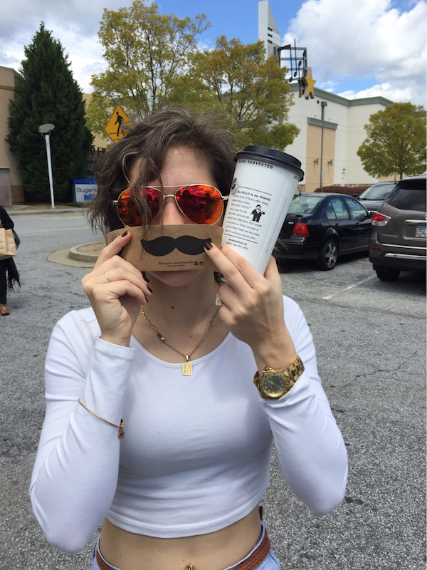 Crazy girl putting fake mustache over lips in white crop top, beautiful golden wrist watch, gold chain around neck, stylish red reflector sunglasses with a white tubler in hand on a road