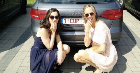girl on left wearing backless halter neck deep blue knee length frock with cool glares and girl on right wearing white lace half sleeves tunic with stylish golden frame sunglasses while sitting togther in the back of Audi car with number plate - 1-CUP