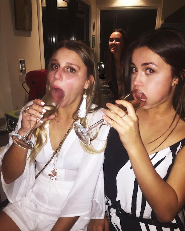 Pretty babes in smart onepiece party outfits inserting wine glass top into their mouth, looking weird with a smiling girl in background