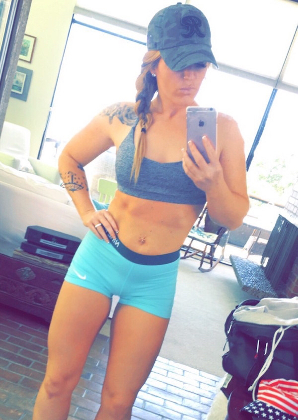 Tattooed girl in a cap, grey sports bra and blue petite pants clicks an iPhone selfie